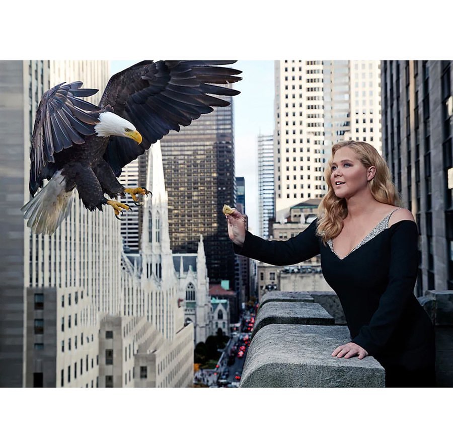 Adult woman outdoors offering food to a bald eagle flying over a city with skyscrapers on a sunny day; beauty shot, fashion style.