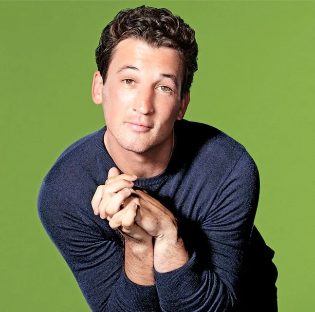 Portrait of actor Miles Teller in a navy sweater against a bright green background, hands clasped, smiling slightly.