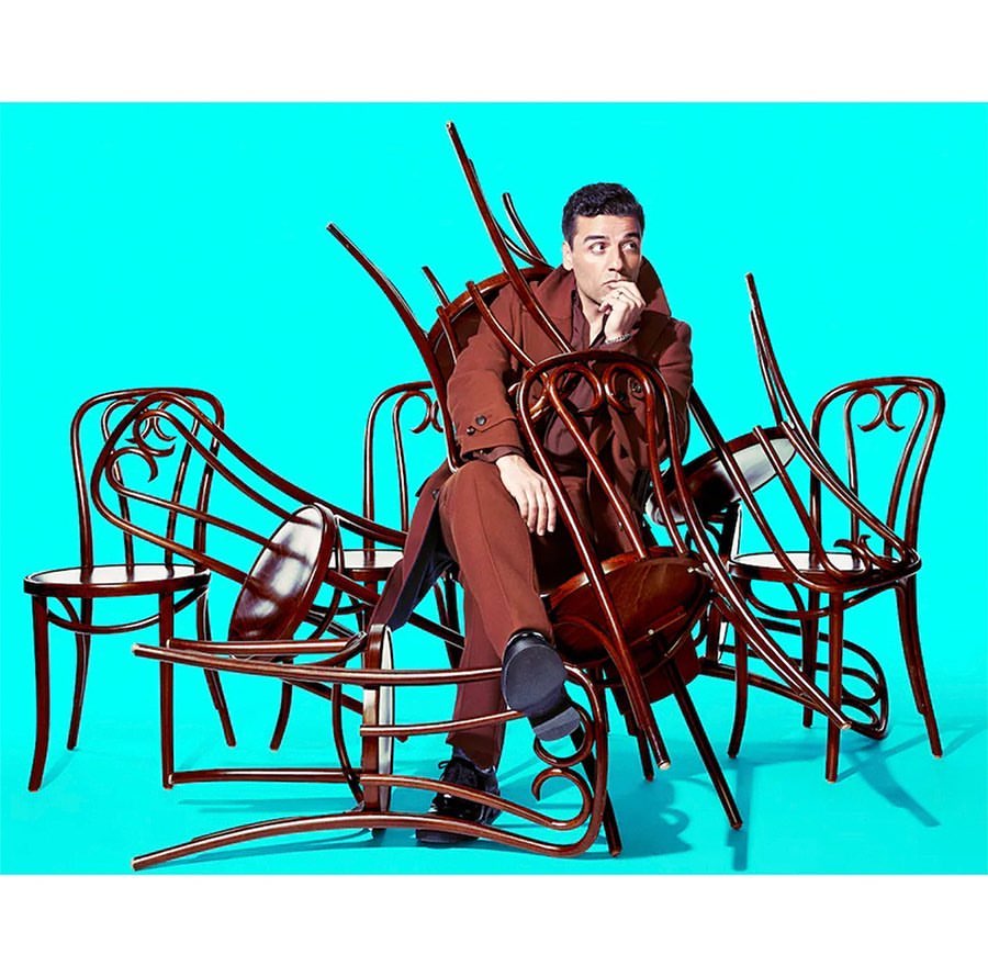 Adult male in a brown suit poses in a pile of bentwood chairs against a teal backdrop in a studio setting with bright lighting.