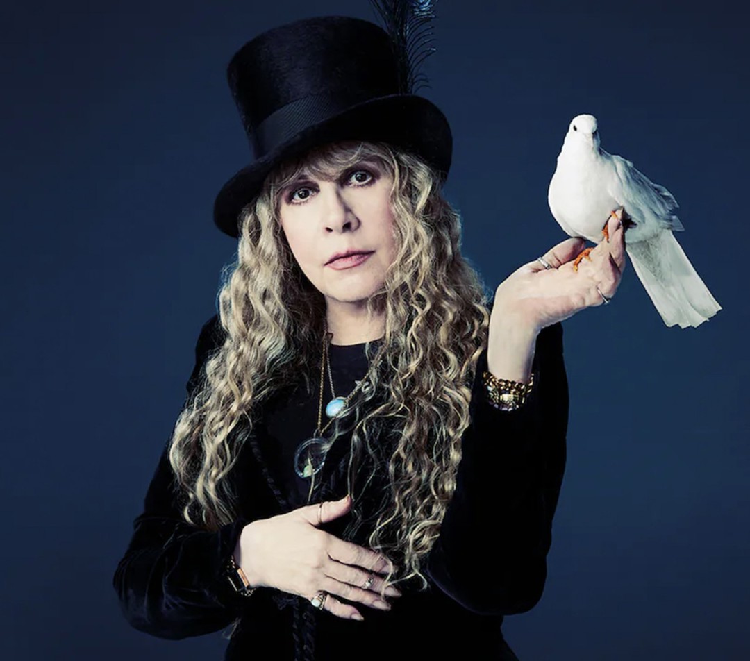 Elegant portrait of a singer with blonde curly hair, wearing a black hat and velvet jacket, holding a white dove on her hand.