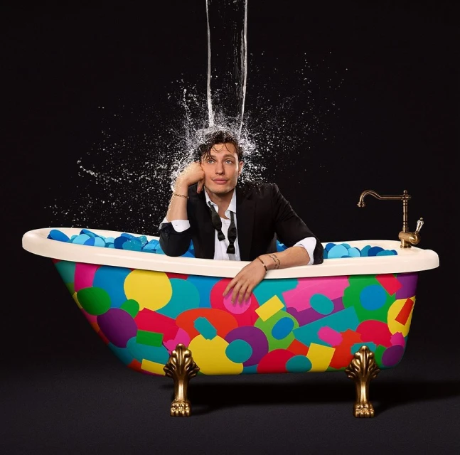 Man in a colourful bathtub is having water poured on him. He is wearing a suit jacket and looks at peace. The background is black.