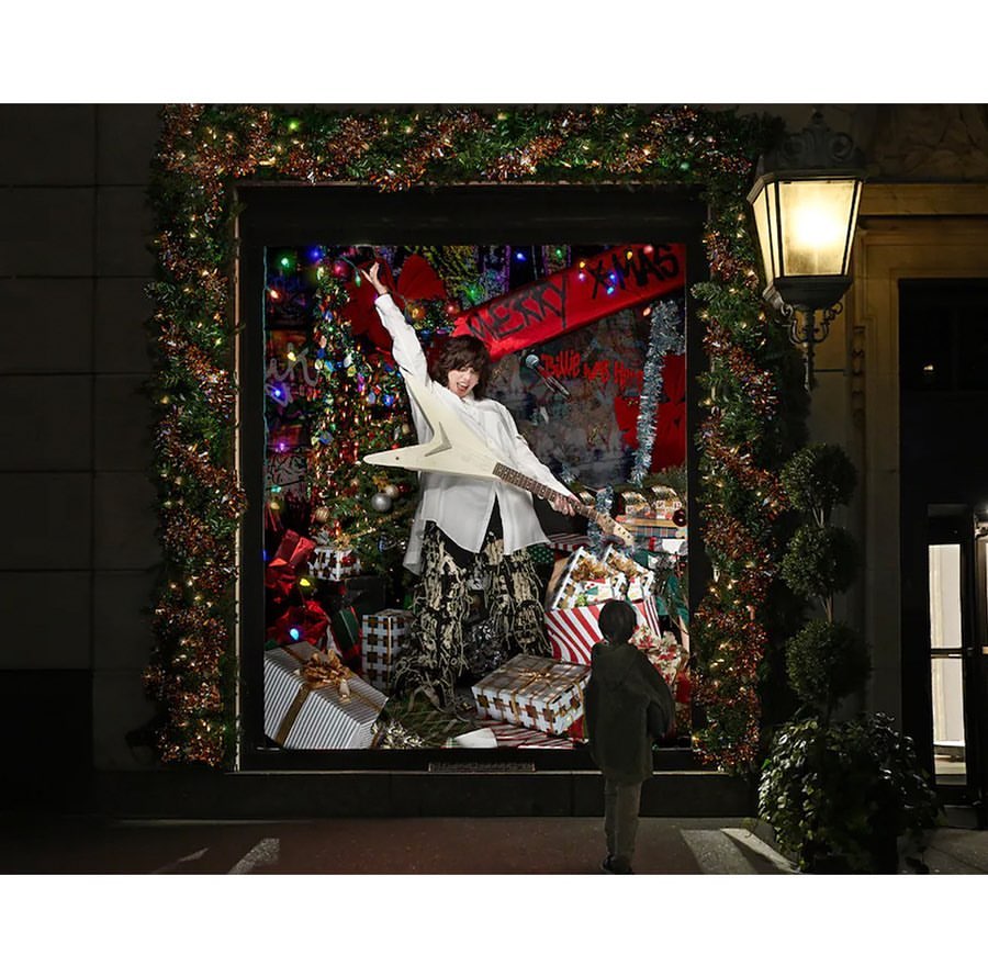 Adult in window display with guitar, surrounded by presents and holiday decor, watched by child. Building exterior visible.