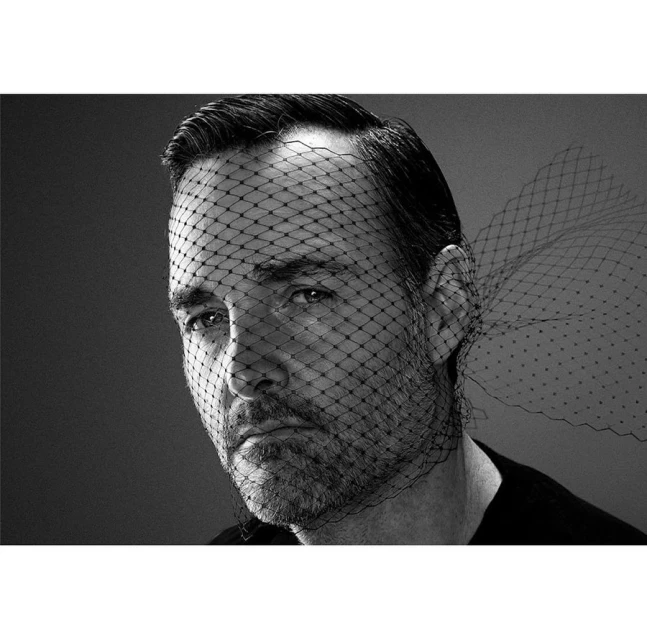 Monochrome portrait of a bearded adult male wearing a black net veil over his face, against a grey backdrop in a studio setting.