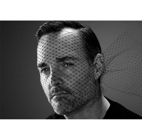 Monochrome portrait of a bearded adult male wearing a black net veil over his face, against a grey backdrop in a studio setting.