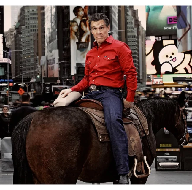 Adult male in a red western shirt and jeans riding a brown horse in an urban setting surrounded by skyscrapers and billboards.