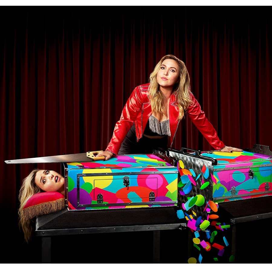 Woman in red jacket poses with colorful magic trick box and saw, with separate head on pillow in front of a dark red stage curtain.