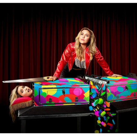 Woman in red jacket poses with colorful magic trick box and saw, with separate head on pillow in front of a dark red stage curtain.