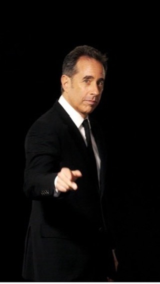 An adult male points his finger directly at the viewer, while wearing a black suit, against a dark black background indoors.