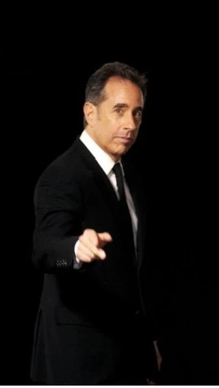 An adult male points his finger directly at the viewer, while wearing a black suit, against a dark black background indoors.