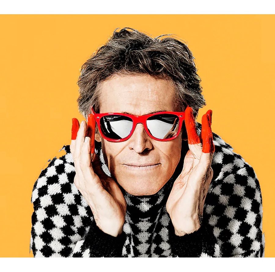 Close-up portrait of a mature adult wearing red glasses and a checker pattern sweater against a solid yellow background, hands raised.