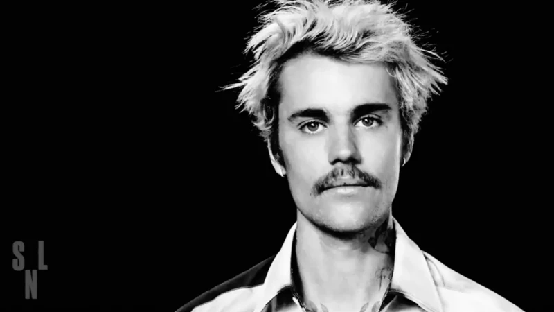 Black and white portrait of Justin Bieber with a mustache, wearing an unbuttoned white shirt, against a dark background.