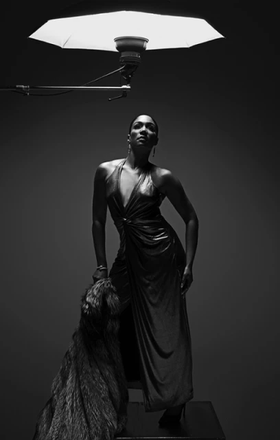 Monochrome studio shot of an adult woman in a gown standing next to a dog, lit by a softbox.