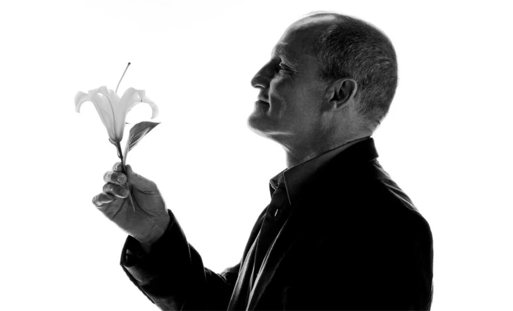 Monochrome portrait of an adult man in a suit holding a white lily flower against a bright white background in a photo studio.