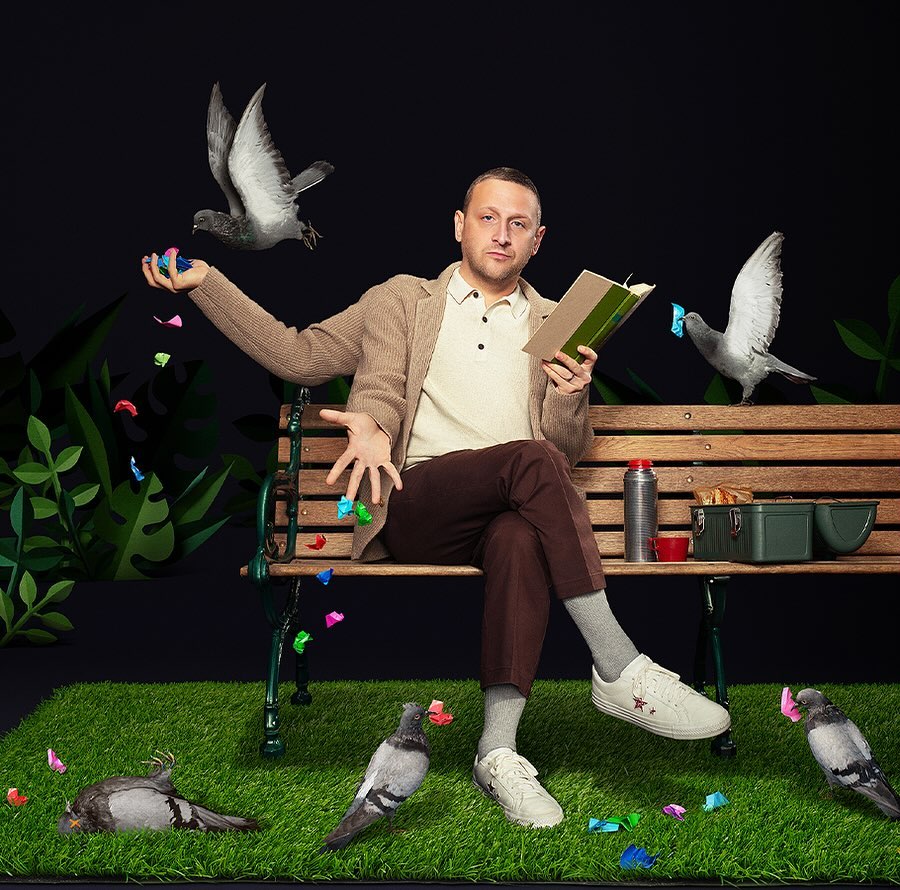 Man in casual attire sits on a bench outdoors surrounded by pigeons and colorful confetti. He is reading a book in front of a dark backdrop.