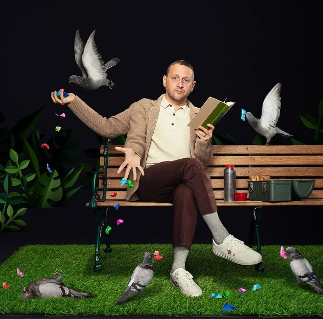 Man in casual attire sits on a bench outdoors surrounded by pigeons and colorful confetti. He is reading a book in front of a dark backdrop.
