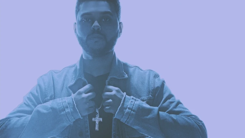 Portrait of an adult male wearing a denim jacket and cross pendant necklace against a light blue background in a studio shot.