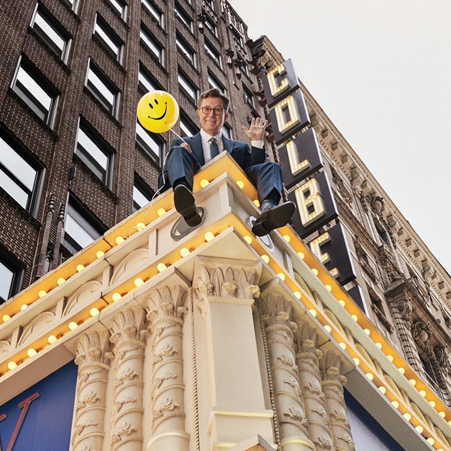 Man in suit sits atop building ledge holding smiley balloon. Urban scene with COLBE signage, architecture, and cloudy sky.