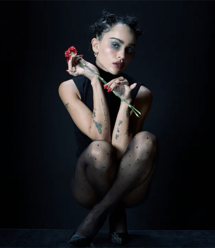 Portrait of a woman with dark hair and a black turtleneck, holding a red flower in her hands, wearing black polka dot tights.