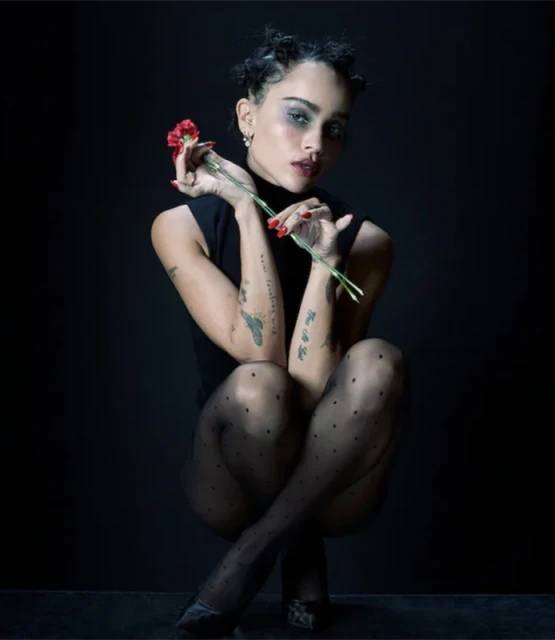 Portrait of a woman with dark hair and a black turtleneck, holding a red flower in her hands, wearing black polka dot tights.