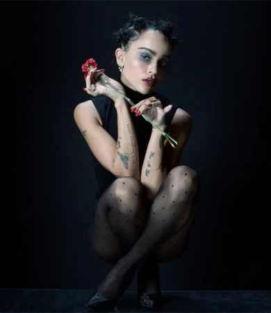 Portrait of a woman with dark hair and a black turtleneck, holding a red flower in her hands, wearing black polka dot tights.