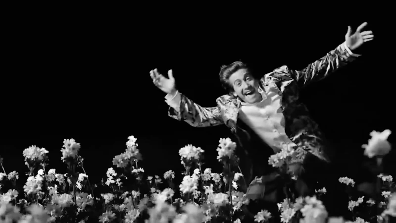 Monochrome image of a man in floral jacket joyfully jumping amidst a field of white flowers against a solid black background.