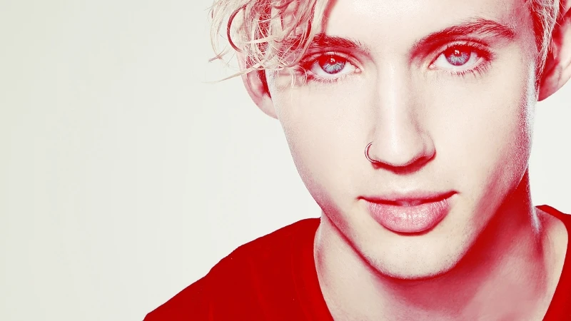 Close-up portrait of a young adult with blond curly hair, blue eyes, and a nose ring, wearing a black shirt against a white backdrop.
