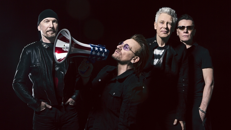 U2 band portrait: Bono holds American flag megaphone, flanked by band members with a dark background. Music and entertainment.