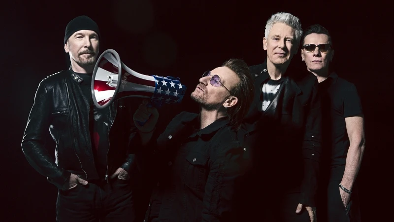 U2 band portrait: Bono holds American flag megaphone, flanked by band members with a dark background. Music and entertainment.