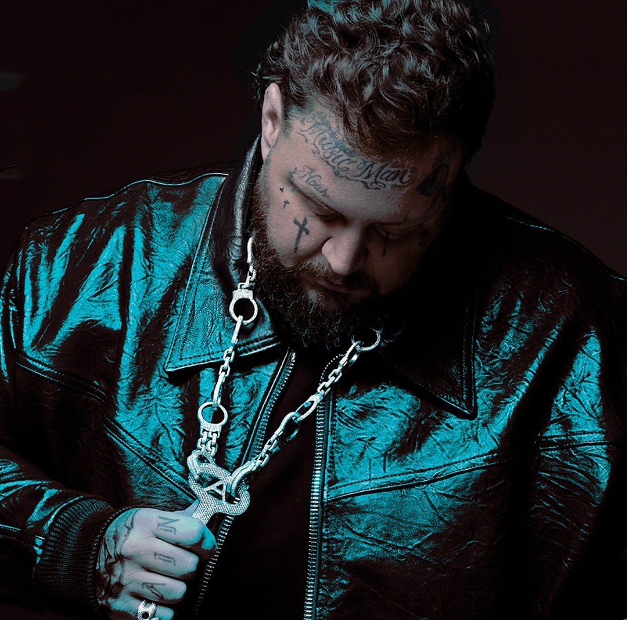 Close-up studio portrait of a tatted man with beard, wearing a black leather jacket and a silver chain with a pendant.