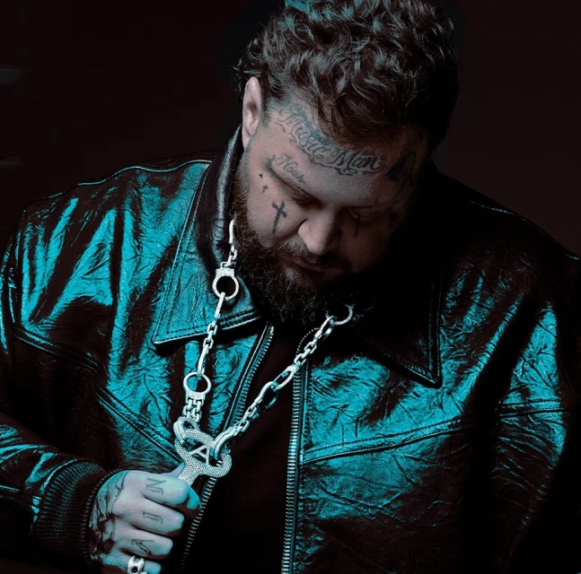 Close-up studio portrait of a tatted man with beard, wearing a black leather jacket and a silver chain with a pendant.