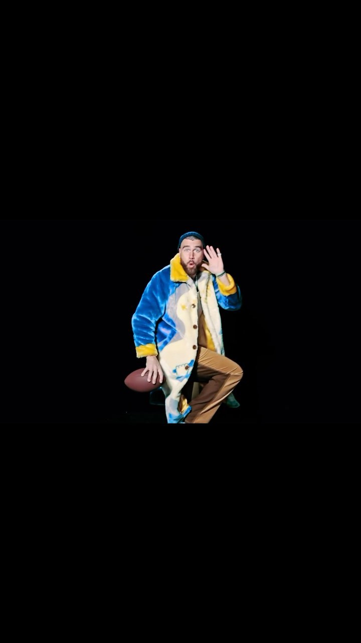 Man posing with football on black backdrop wearing blue, yellow and beige coat, tan pants, waving his hand as if giving a greeting.