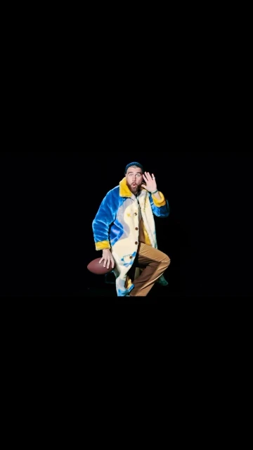 Man posing with football on black backdrop wearing blue, yellow and beige coat, tan pants, waving his hand as if giving a greeting.