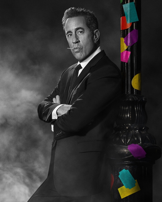 Jerry Seinfeld in a black suit and toothpick, leaning against a polka-dotted pole in a black and white portrait against a smoky background.