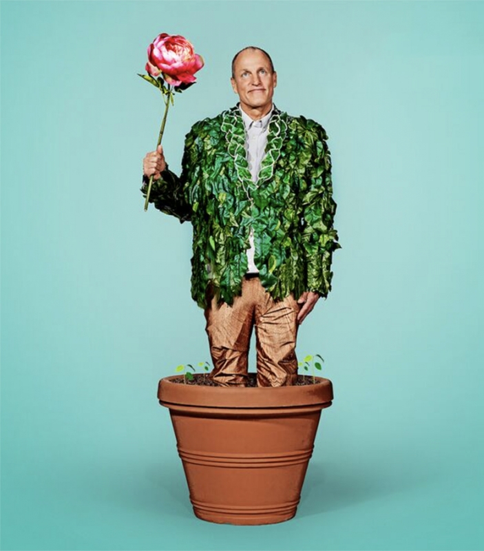Woody Harrelson standing inside a large terracotta flower pot wearing a leaf jacket and holding a large pink flower.