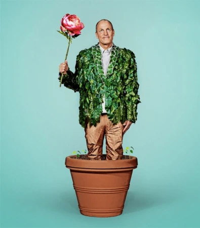Woody Harrelson standing inside a large terracotta flower pot wearing a leaf jacket and holding a large pink flower.