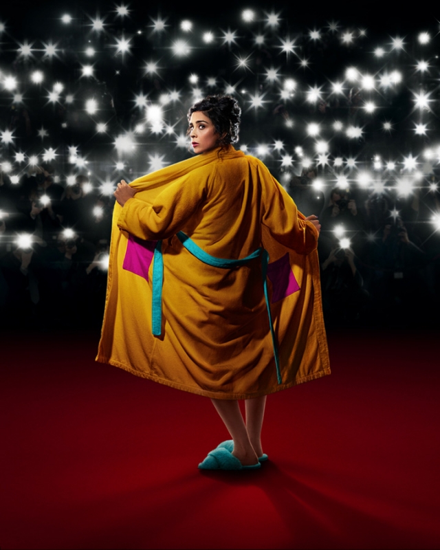 Woman in a gold robe and blue slippers standing on a red carpet in front of a backdrop of bright stage lights, looking over her shoulder.
