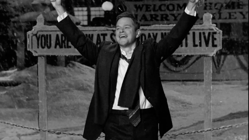 Monochrome image features a smiling man with arms raised outside, with snow falling near a 'Welcome Home Harry Bailey' sign.