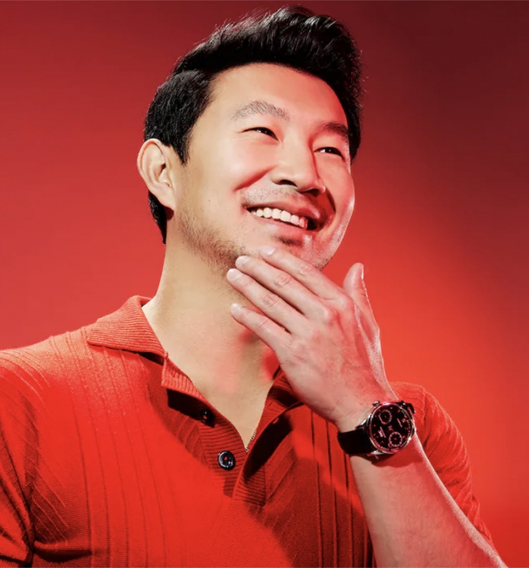 Simu Liu smiles broadly in a red polo shirt, his hand gently touching his chin, a watch visible on his wrist against a red backdrop.