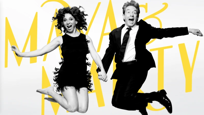 Two performers, a man and a woman, in formal attire joyfully leap mid-air against a bright, simple background with stylized yellow text.