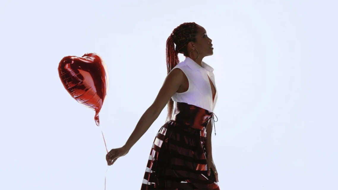 An adult woman in a white blouse and red skirt poses in front of a white background while holding a red heart-shaped balloon.