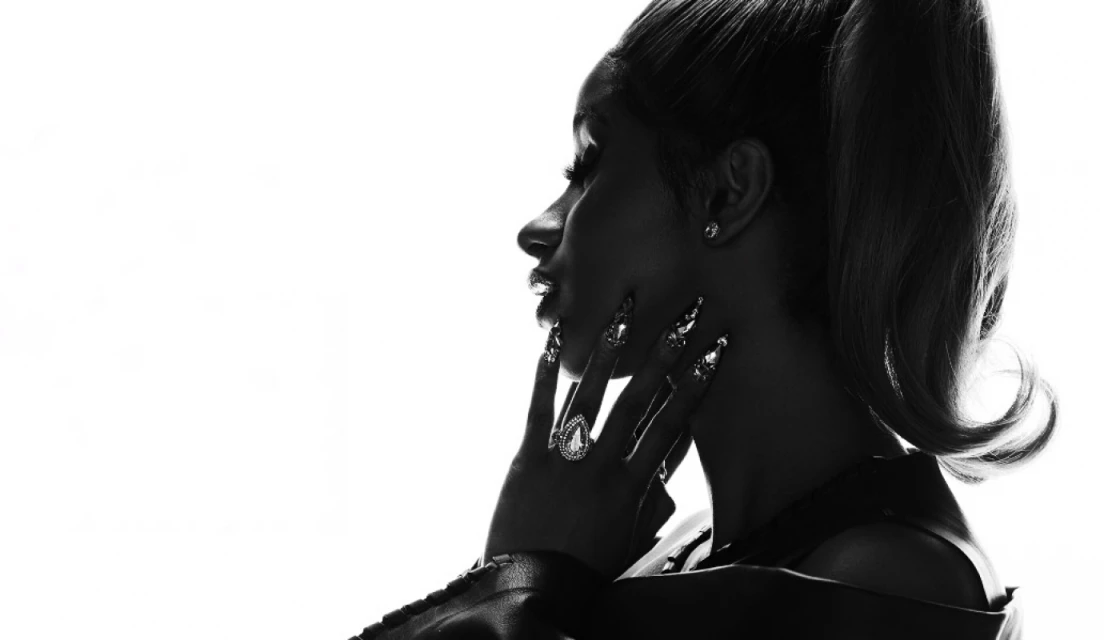 Monochrome portrait of an adult female with a high ponytail, diamond earrings, bejeweled nails and a ring, placing her hand on her neck.
