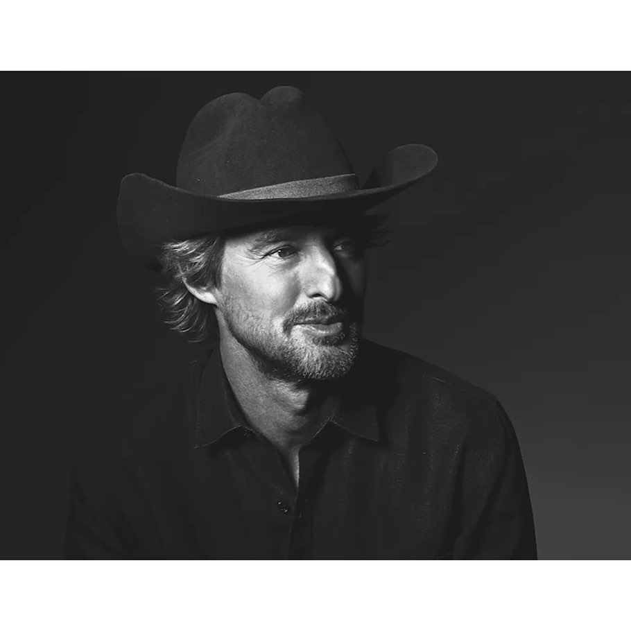 Monochrome portrait of a smiling adult male wearing a cowboy hat and collared shirt against a gradient dark background.