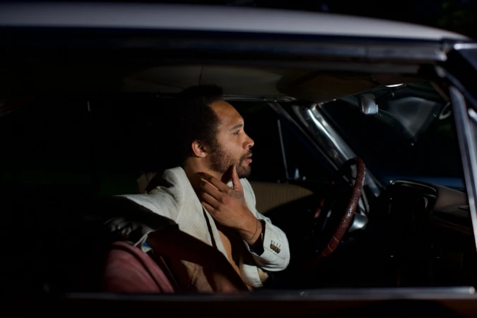 A man sitting inside a car at night, touching his chin with one hand and wearing a white jacket.