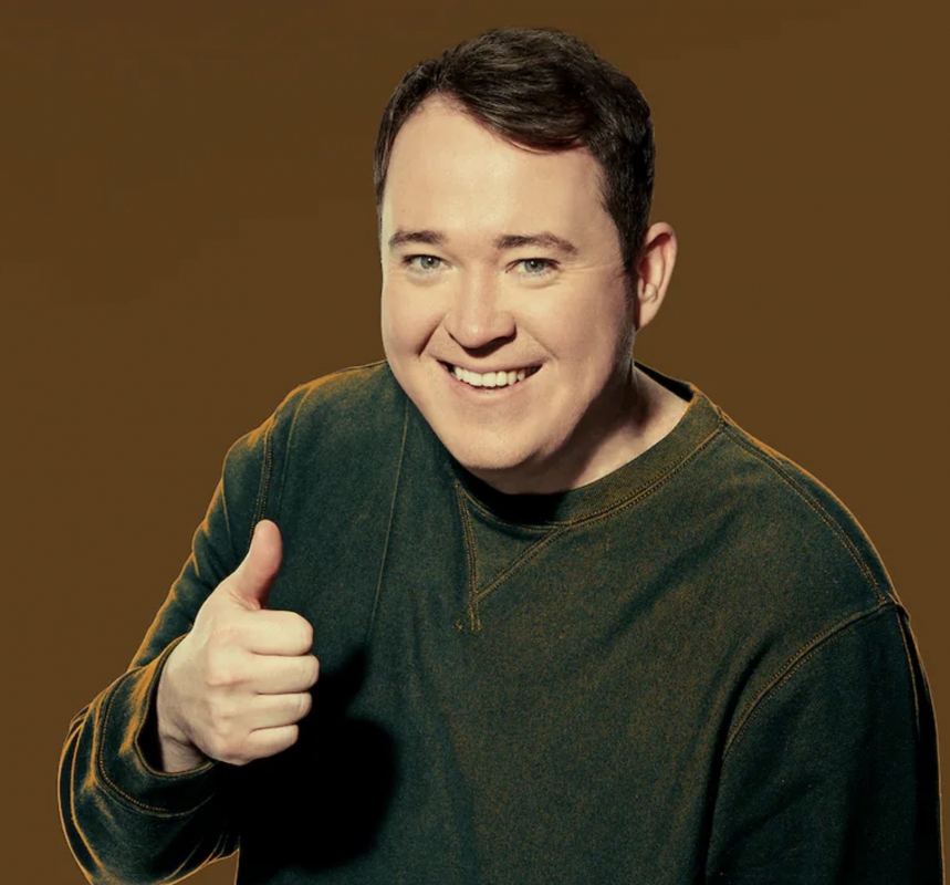 Portrait of Shane Gillis wearing a dark olive green crewneck sweatshirt giving a thumbs up against an olive background.