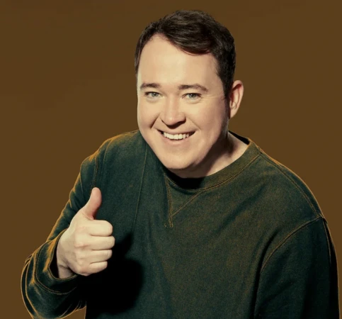 Portrait of Shane Gillis wearing a dark olive green crewneck sweatshirt giving a thumbs up against an olive background.