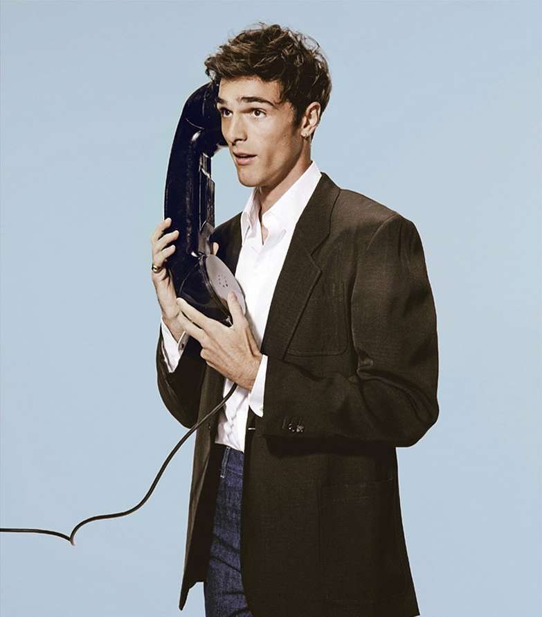 Man holds vintage telephone receiver to his ear, wearing a suit jacket and collared shirt, posing in a light blue setting.