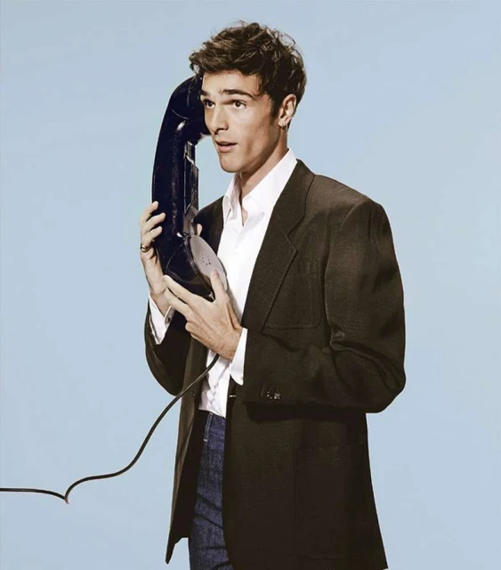 Man holds vintage telephone receiver to his ear, wearing a suit jacket and collared shirt, posing in a light blue setting.