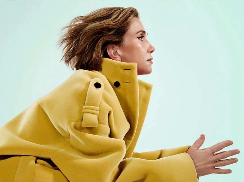 Woman in yellow coat poses in profile against a light blue background. Fashion editorial shot with focus on the coat's design.