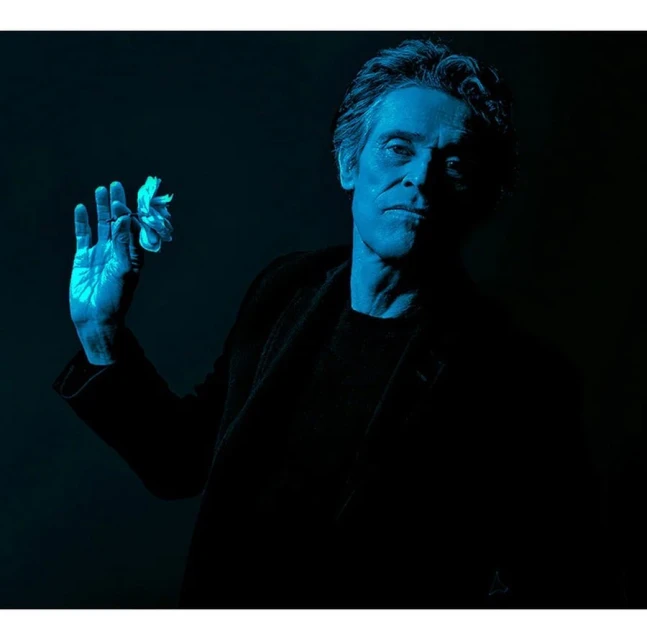 Portrait of an adult man with gray hair in a black coat, illuminated by blue light and holding a bright white flower in his hand.