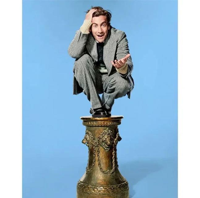 Jake Gyllenhaal in a crouched pose on an ornate pedestal against a solid blue background, wearing a grey suit and sweater.